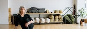 Older woman sitting on a yoga mat in a bright home studio, smiling and preparing for gentle exercise.