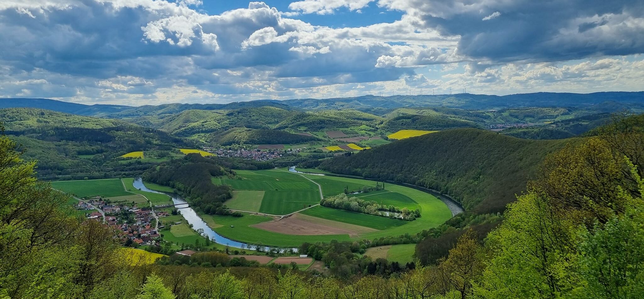 A winding river curves through green fields and villages beneath wooded hills and a cloud-filled sky.